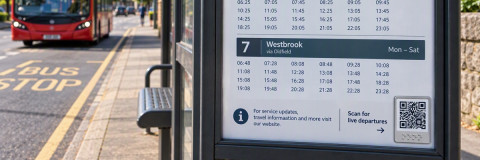 a bus stop timetable on a bus shelter with a QR code with a bus in the distance.